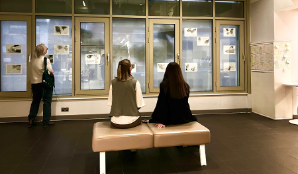 Two people sit on a bench at an exhibition and look at art work on the walls