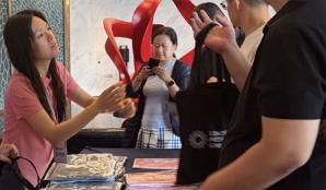 A woman hands a London Met branded black tote bag to a person at a desk, there are people behind